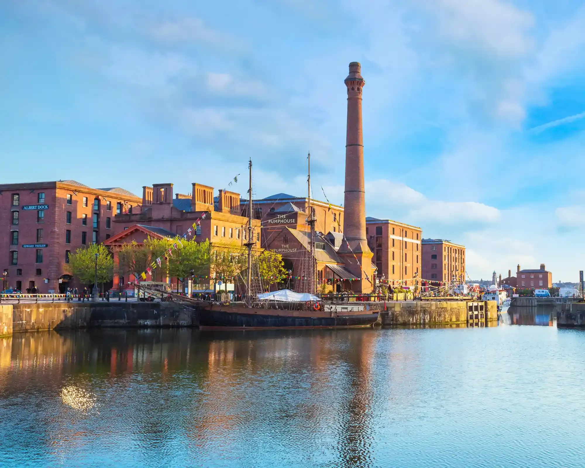 Albert Dock in Liverpool on a sunny day, with the Pumphouse chimney, red-brick warehouses and a tall ship reflected in the water.