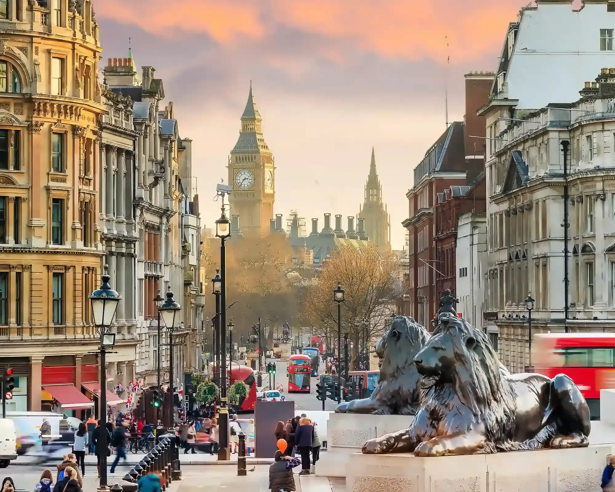 View from Trafalgar Square towards Big Ben at sunset, with a bronze lion statue and red buses on Whitehall.