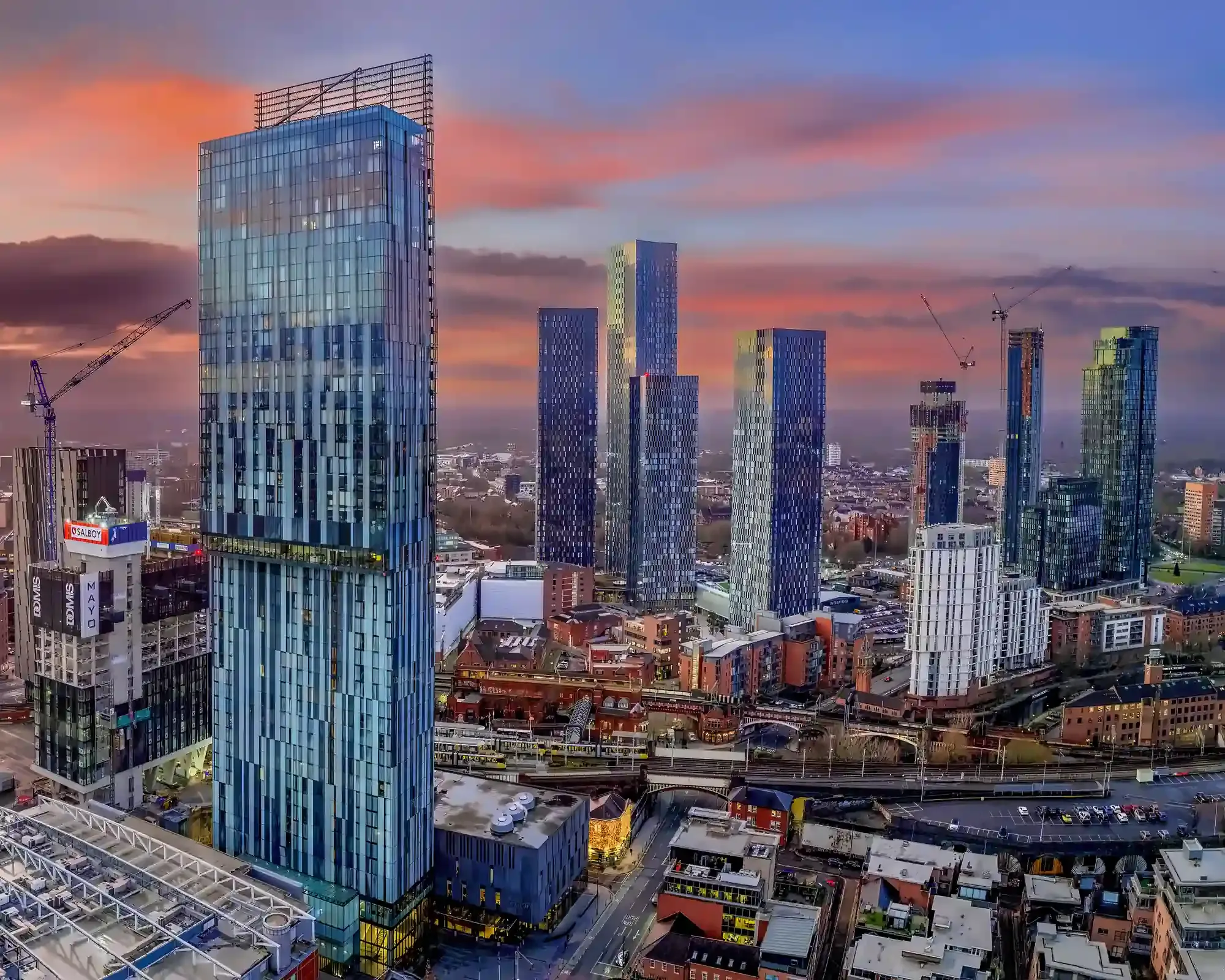 Manchester skyline at sunset, with the glass Beetham Tower in front of the Deansgate Square skyscrapers under a pink and orange sky.