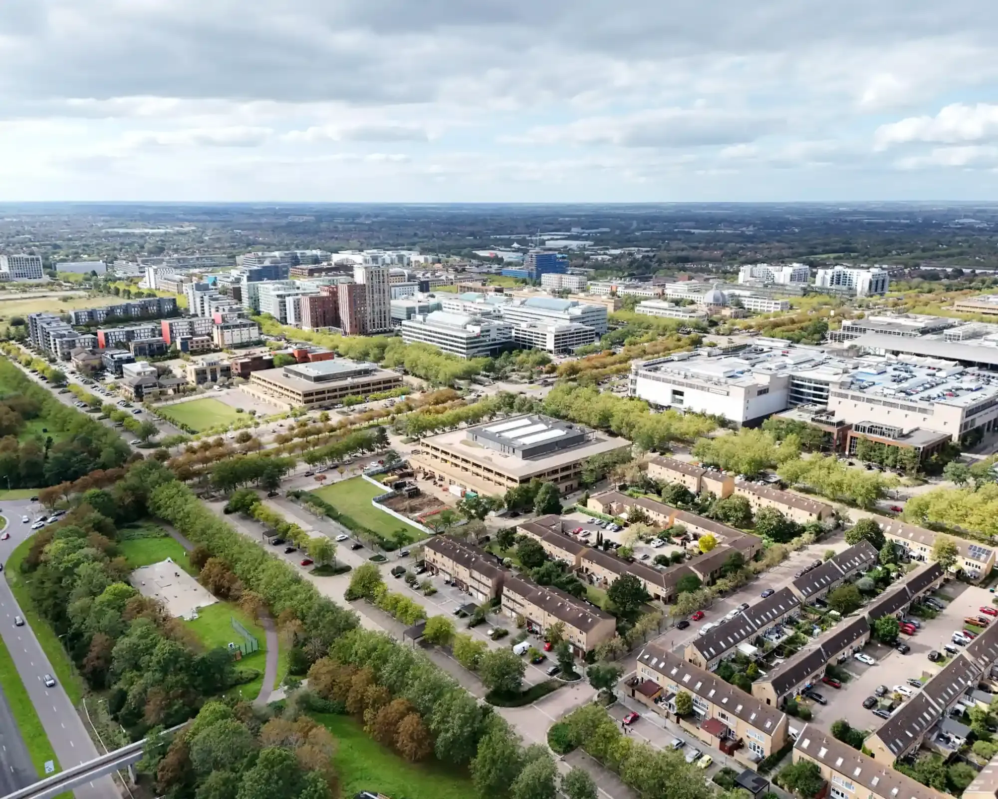 Aerial view of central Milton Keynes, with tree-lined boulevards, office blocks and the shopping centre stretching towards green countryside under a cloudy sky.