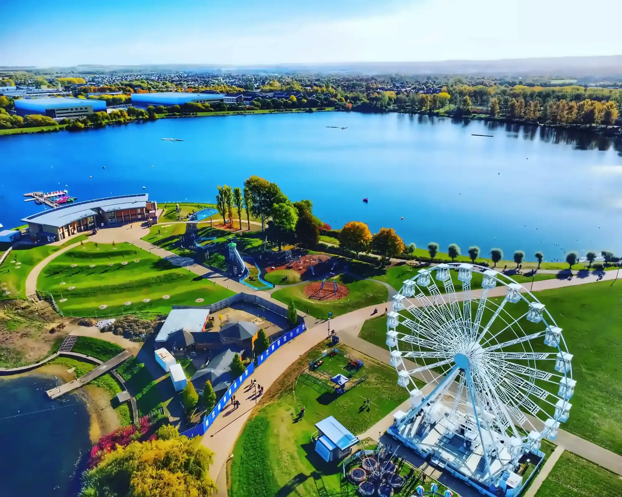 Aerial view of Willen Lake in Milton Keynes on a sunny autumn day, with a big wheel, a playground and green parkland beside the water.