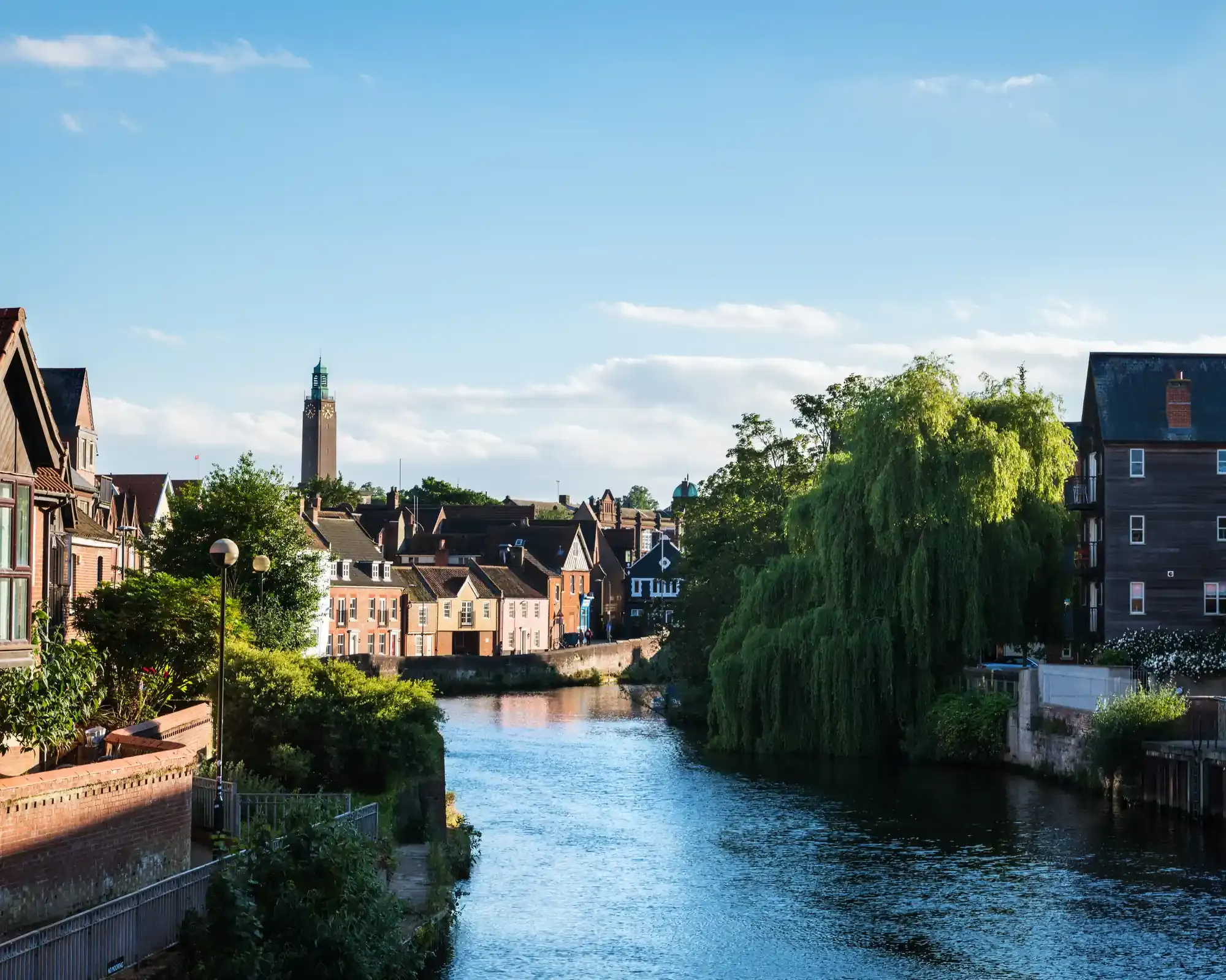 The River Wensum in Norwich on a sunny evening, with riverside houses, a weeping willow and City Hall's clock tower in the distance.