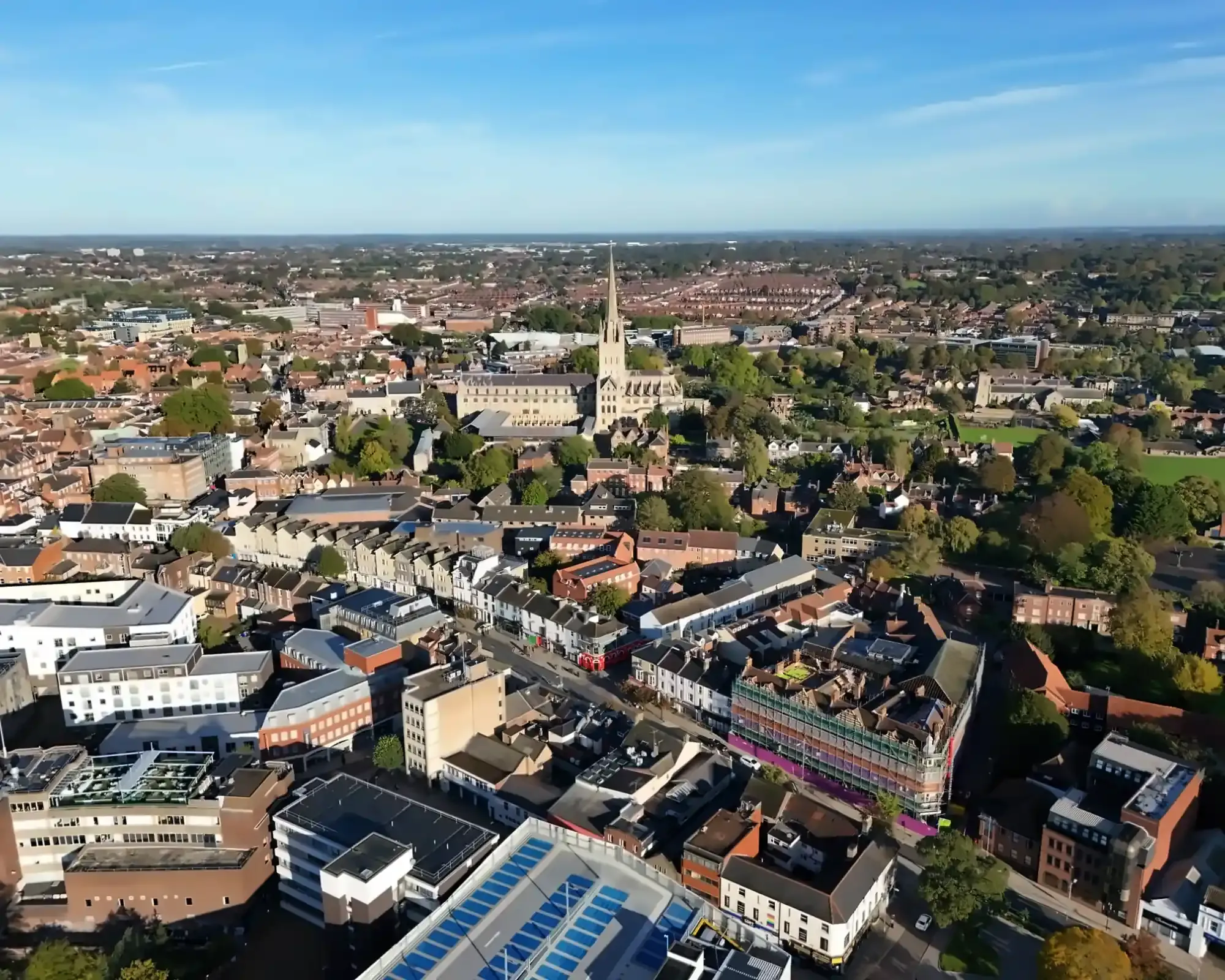 Aerial view of Norwich on a sunny day, with the cathedral's spire rising above red-brick rooftops and the city centre streets.