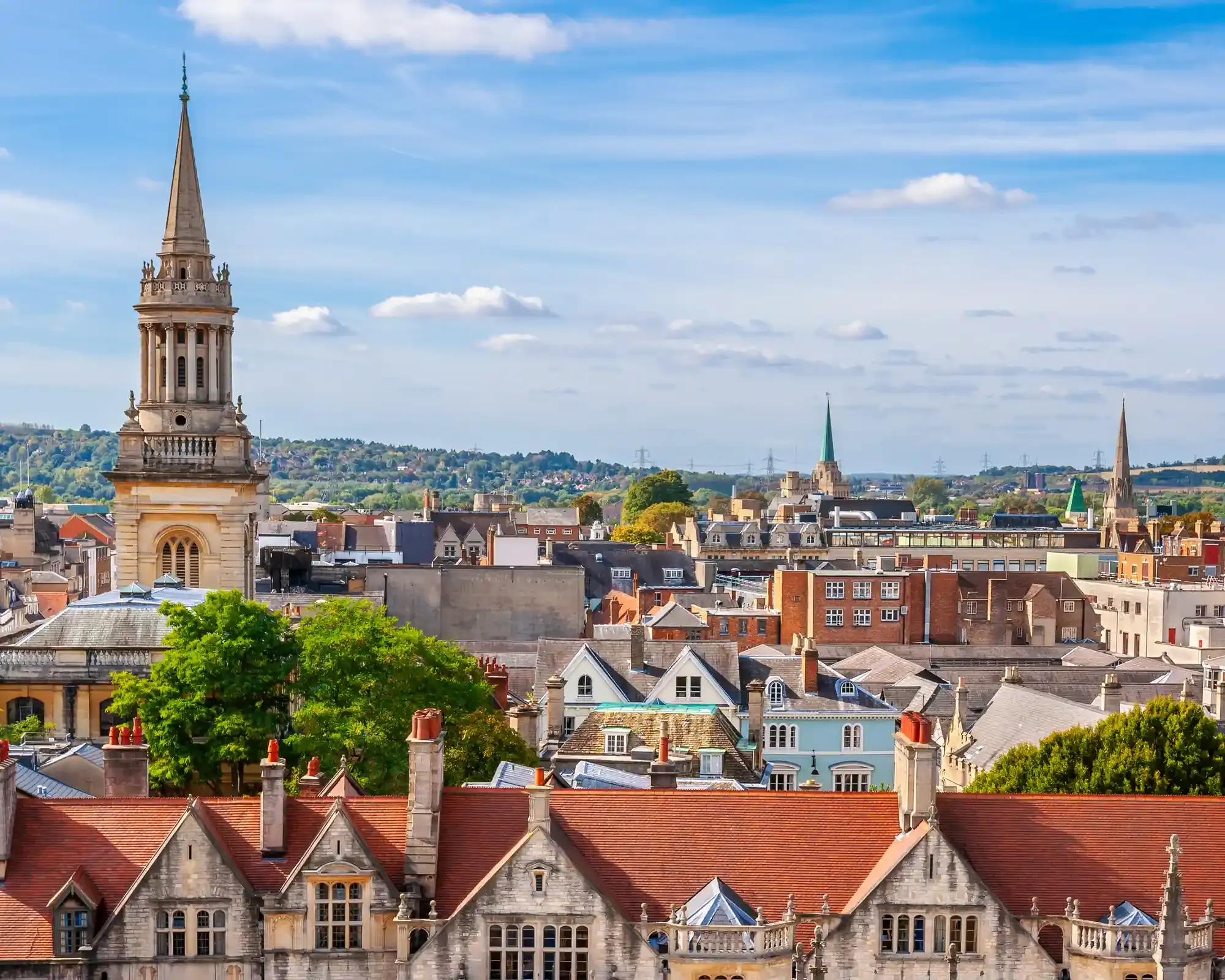 Oxford's rooftops on a sunny day, with the spire of Lincoln College Library rising above stone gables, red-tiled roofs and distant green hills.