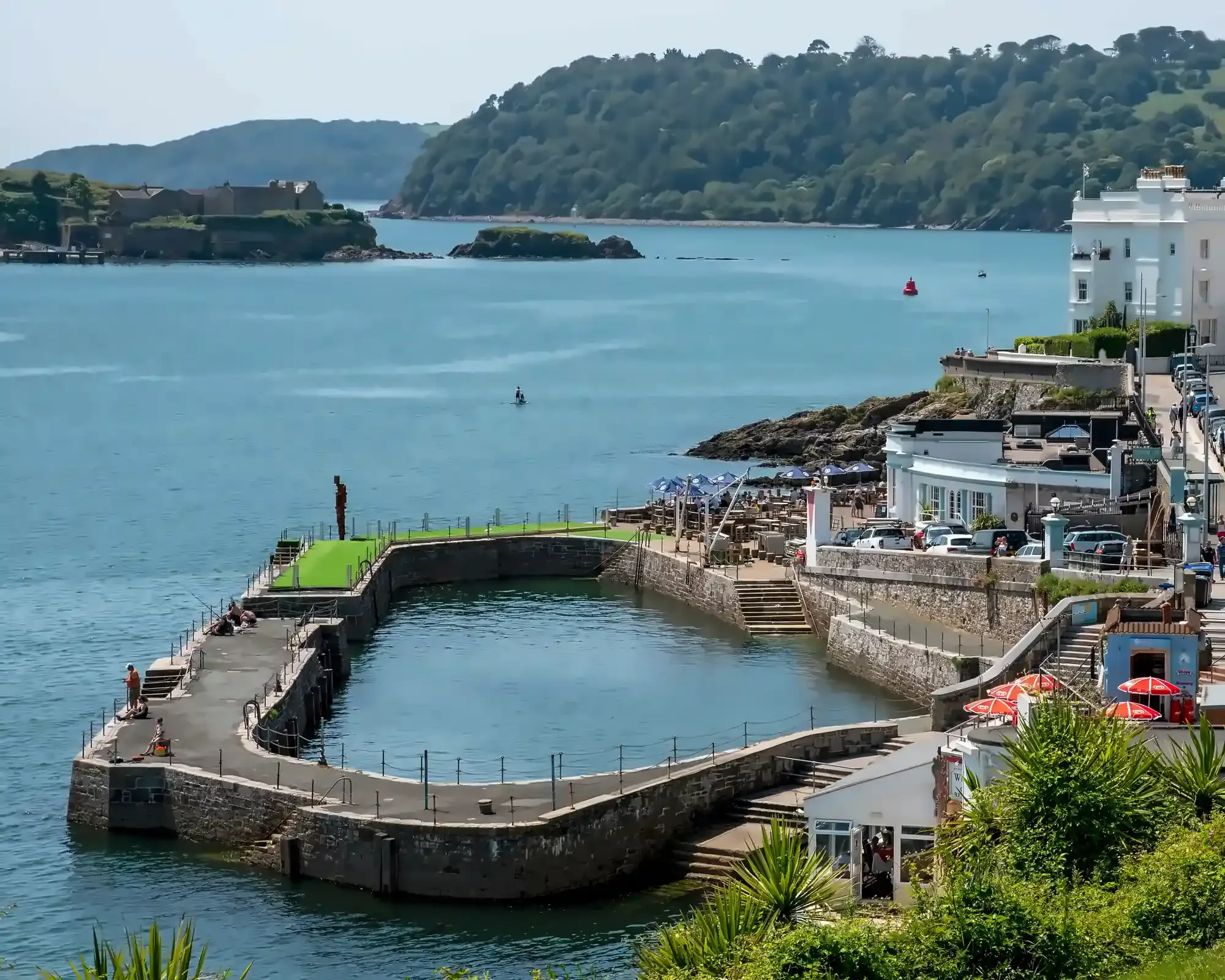 Plymouth's waterfront on a sunny day, with a stone-walled tidal pool beside Plymouth Sound, and Drake's Island and wooded hills across the water.