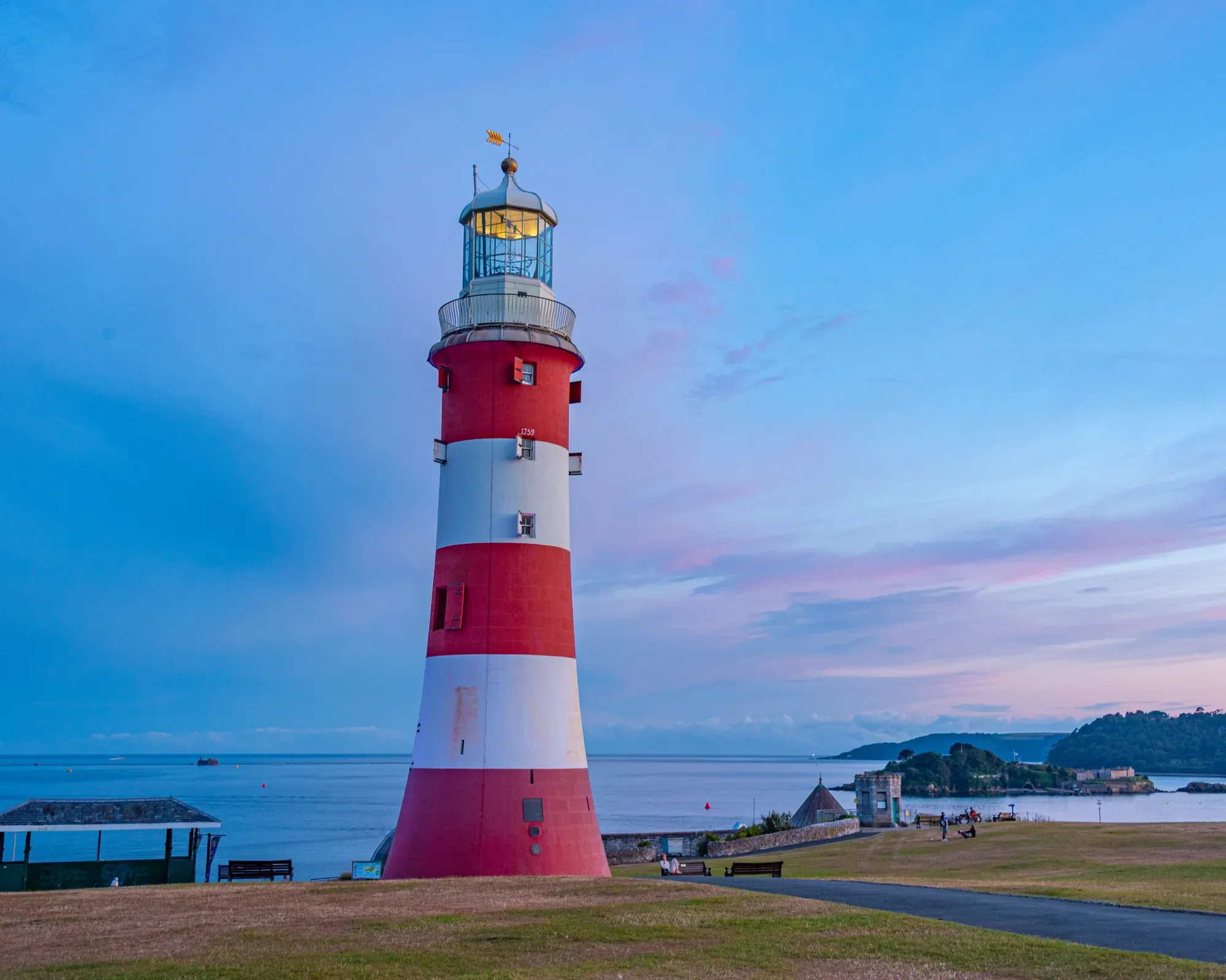 Smeaton's Tower, the red and white striped lighthouse on Plymouth Hoe, at dusk, with Plymouth Sound and Drake's Island behind under a pink and blue sky.