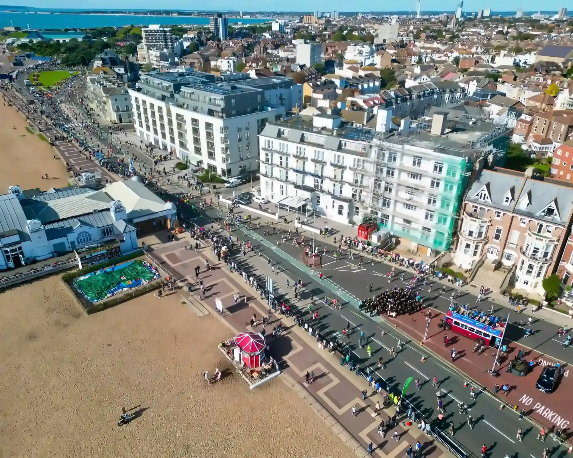 Aerial view of Southsea seafront in Portsmouth on a sunny day, with runners and crowds along the road beside the shingle beach, and the Spinnaker Tower on the skyline beyond.