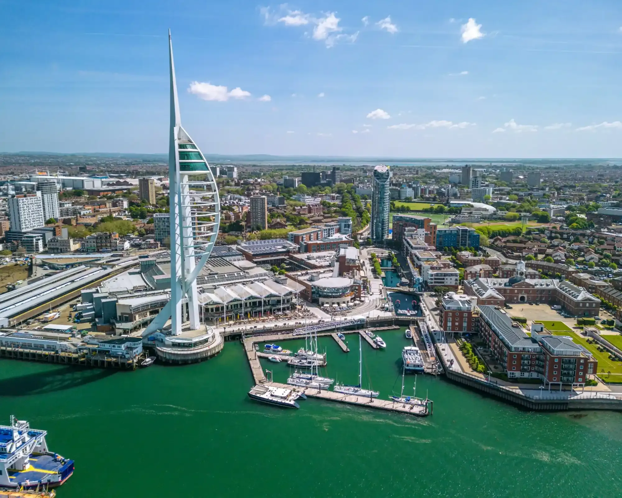 Aerial view of the Spinnaker Tower and Gunwharf Quays in Portsmouth on a sunny day, with yachts moored in the marina and the city beyond.
