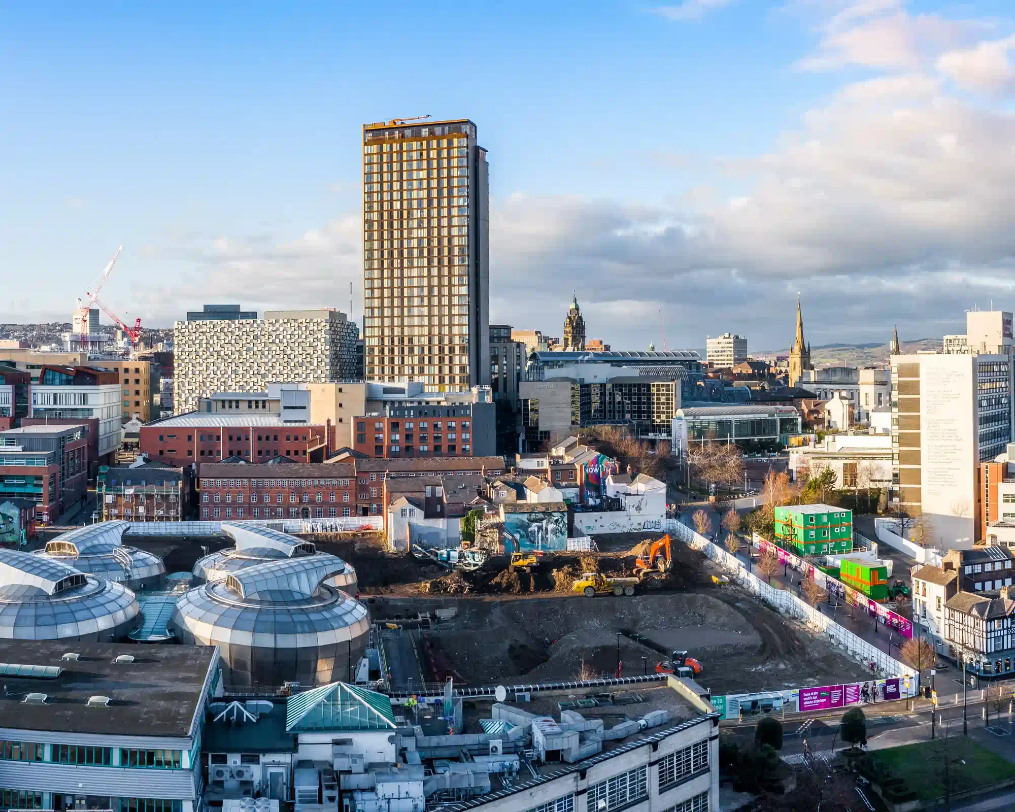 Sheffield city centre on a sunny day, with a tall tower block rising above domed glass roofs, red-brick buildings and the Town Hall clock tower, and green hills beyond.