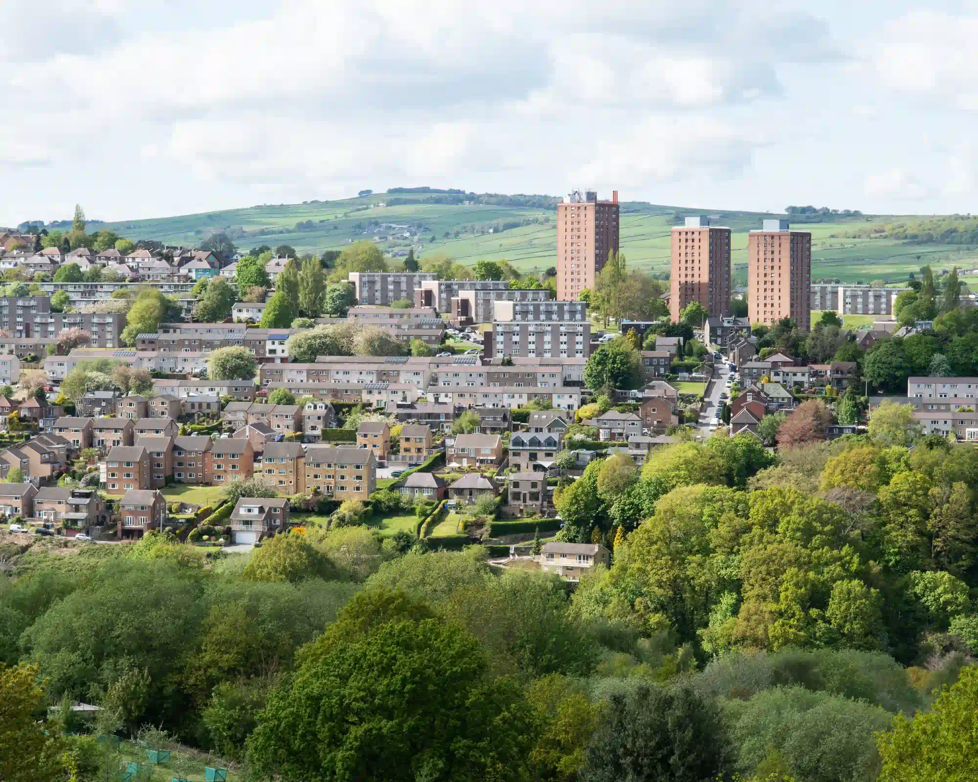 A hillside suburb of Sheffield with rows of houses and three red-brick tower blocks, surrounded by leafy trees, with green fields rising behind.
