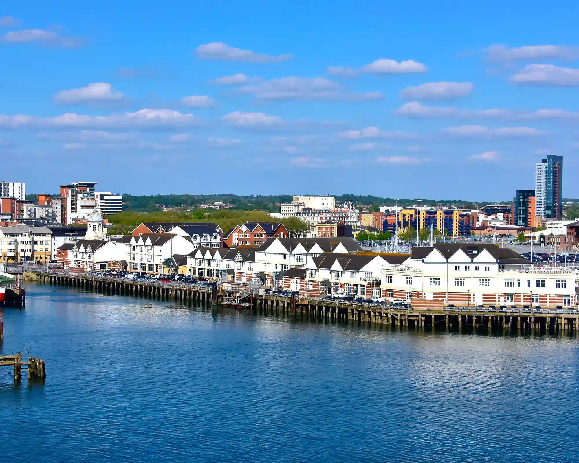 Southampton's Town Quay on a sunny day, with white waterfront buildings along a wooden pier, a ferry at its berth and the city skyline under a blue sky.