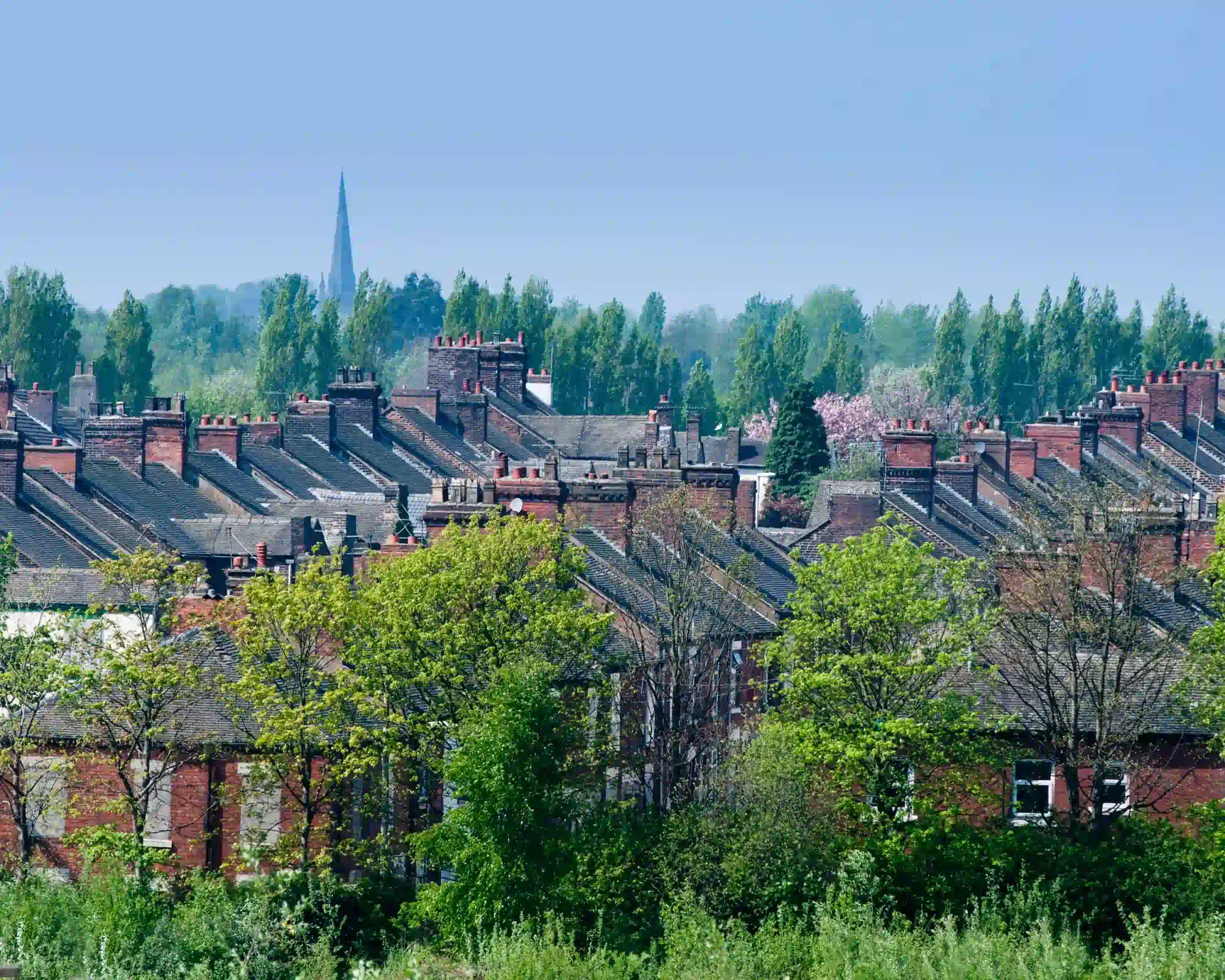 Rows of red-brick terraced houses with chimney pots in Stoke-on-Trent, surrounded by green trees, with a church spire rising in the distance under a blue sky.