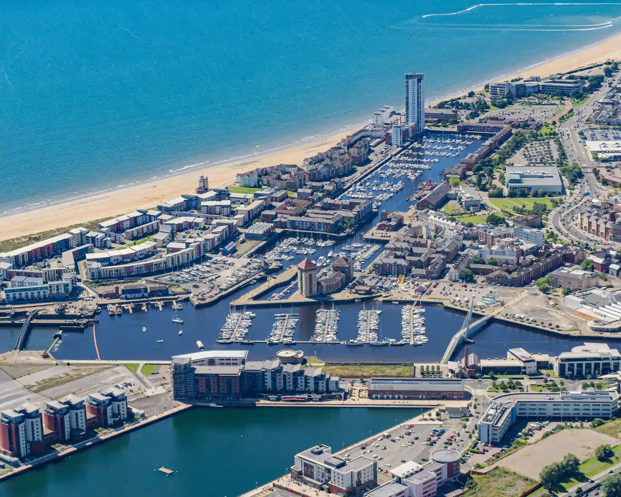 Aerial view of Swansea Marina and the Maritime Quarter on a sunny day, with rows of moored boats, the Meridian Tower and a sandy beach curving along the blue sea.