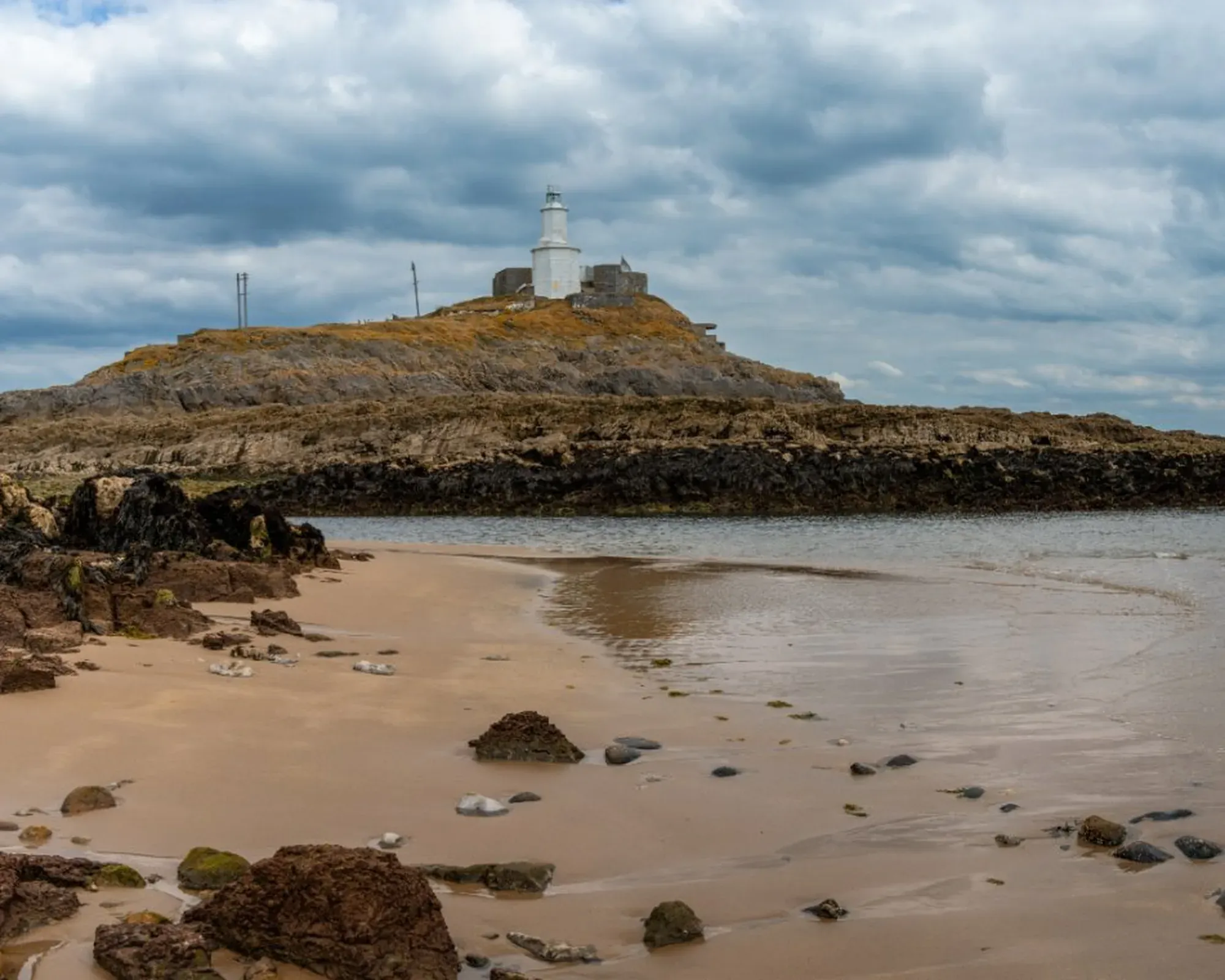Mumbles Lighthouse on its rocky headland near Swansea, seen from a sandy beach scattered with stones, under a cloudy sky.