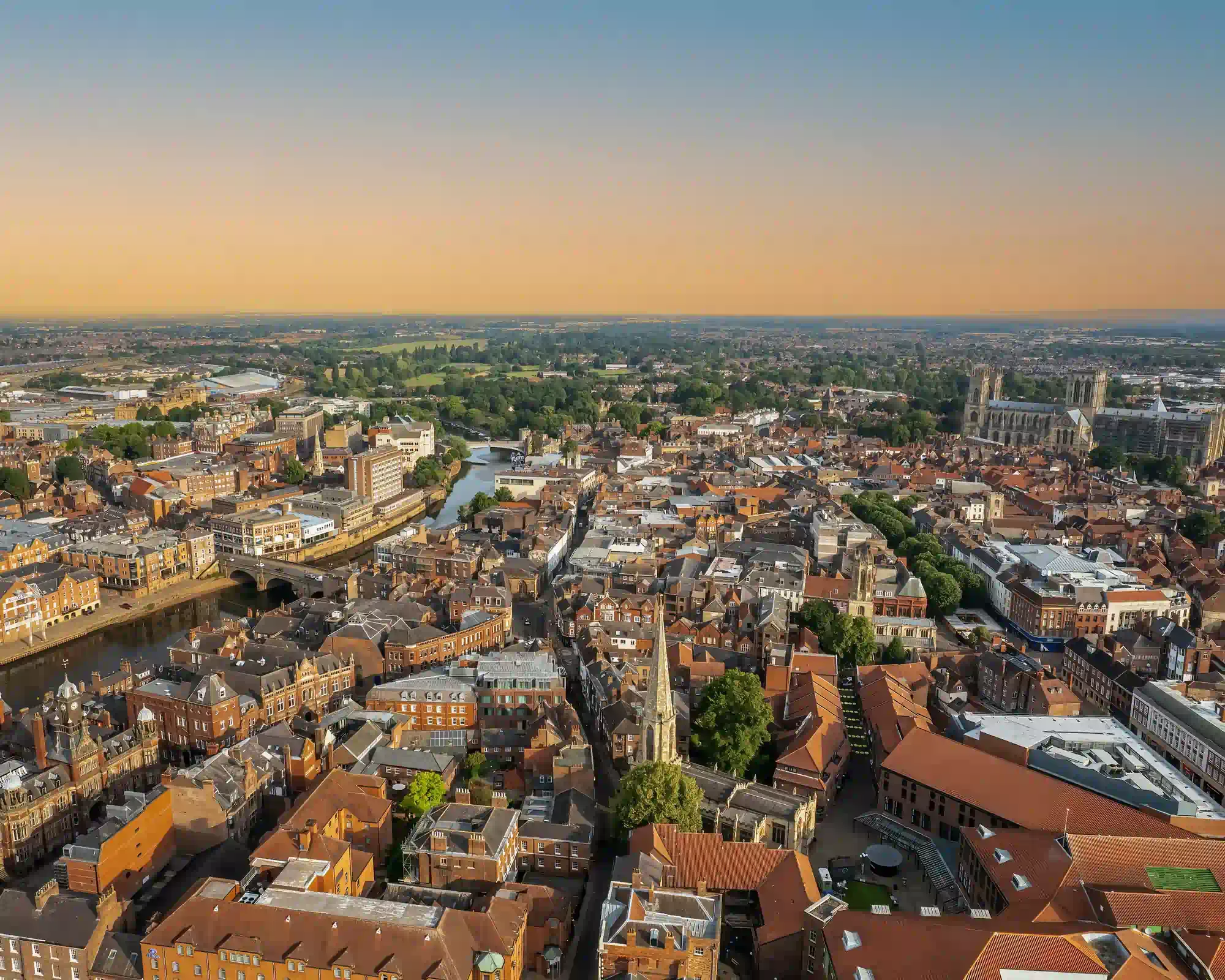 Aerial view of York at sunset, with the River Ouse winding through red-roofed streets towards York Minster, under an orange and blue sky.