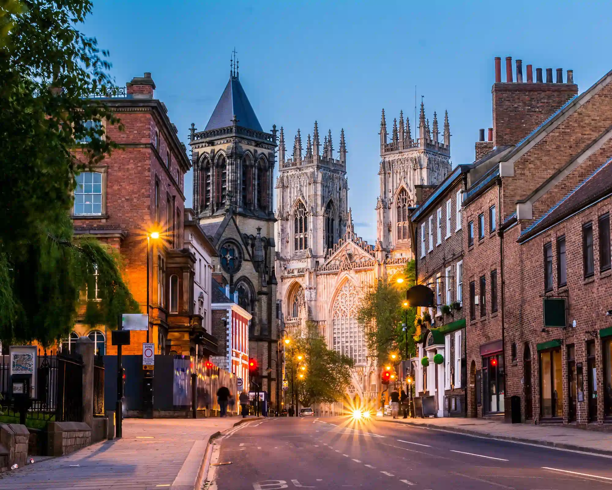 York Minster lit up at dusk, seen along a quiet street lined with red-brick buildings and glowing streetlights.